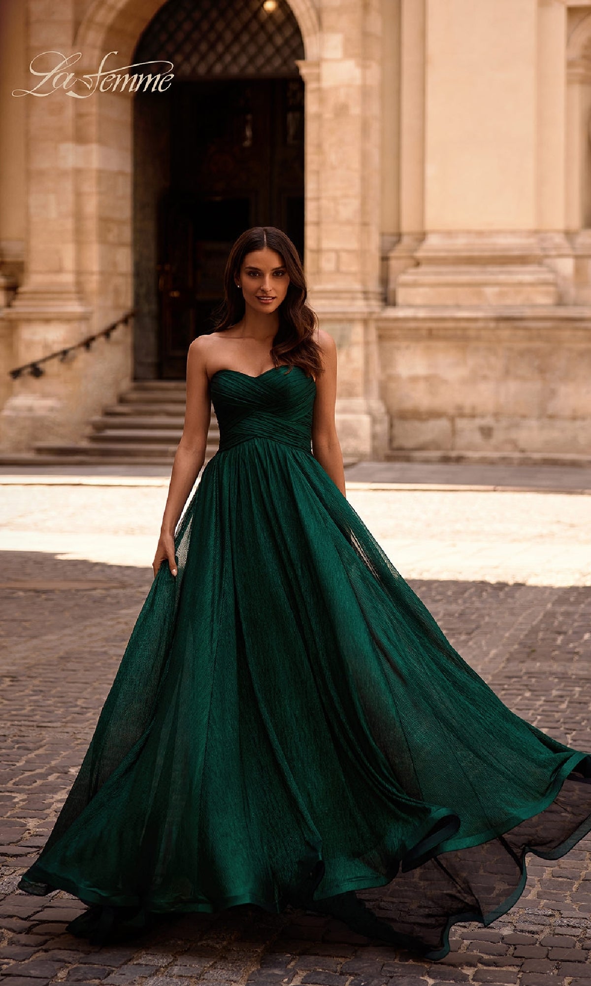 A woman in a strapless long metallic La Femme 32382 prom dress stands on a cobblestone street before an ornate stone building.